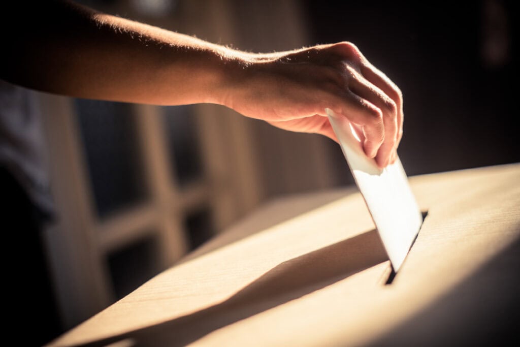 A hand is inserting a piece of paper into a slot on a wooden surface, possibly casting a vote or submitting a paper.