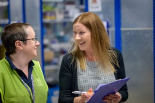 Two smiling women speak to each other in a warehouse. One holds a clipboard and pen.