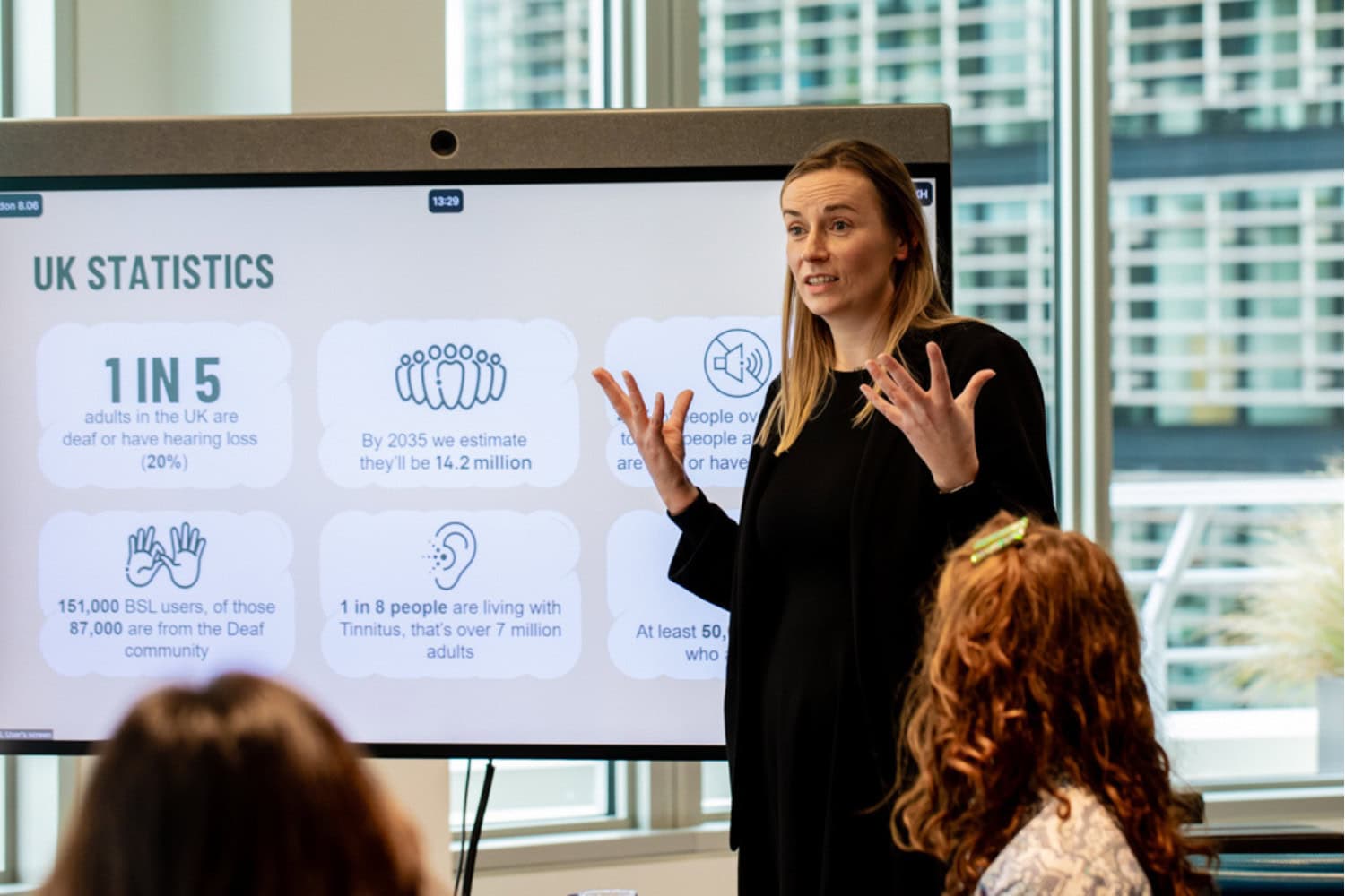 A woman stands beside a big screen presenting. The screen displays a PowerPoint, with statistics about hearing loss in the UK.