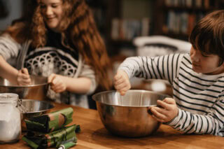 Two children sit at a table with silver mixing bowls, mixing cake mix.
