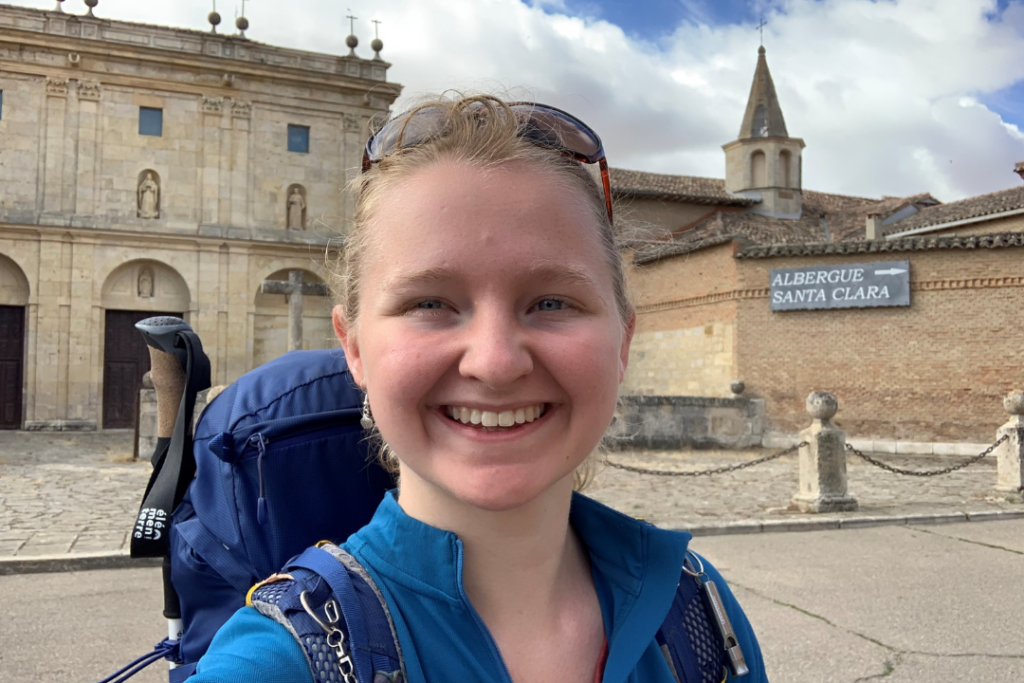 Mary, a young woman wearing a large rucksack with hiking poles, stands and smiles for the camera outside of a European building. A sign in the background reads: "Albergue Santa Clara".