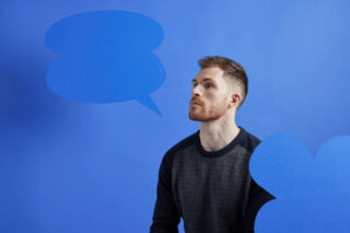 A man wearing a black jumper sits in front of a blue background, looking off to the side. He wears a hearing aid, There are blue speech bubble icons next to hope.