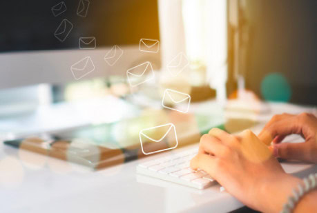 A person's hands over a computer keyboard in front of a screen. There are envelope icons overlaying the image representing the sending of emails.