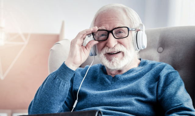 Elderley man seated in an armchair, wearing glasses and headphones, holding a tablet device and smiling at the camera.