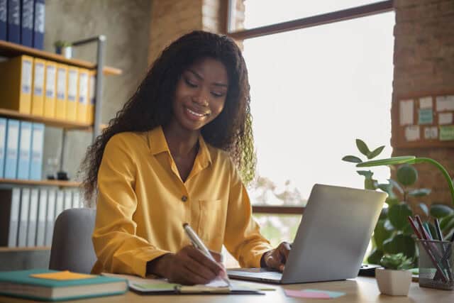 A woman sits at a desk in an office with a laptop, writing into a notepad. She is wearing a yellow shirt and smiling.