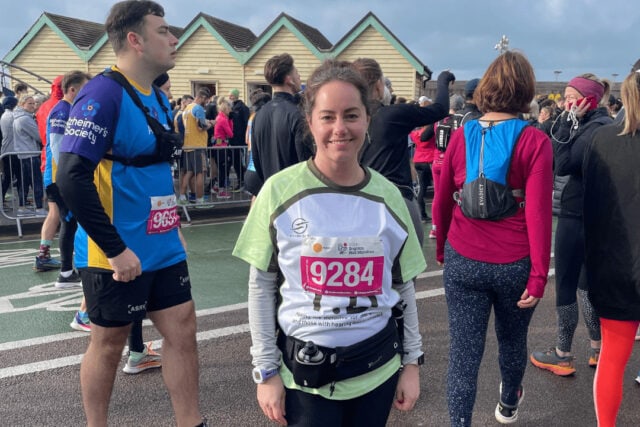 A woman stands with other runners. She wears a running number and is smiling.