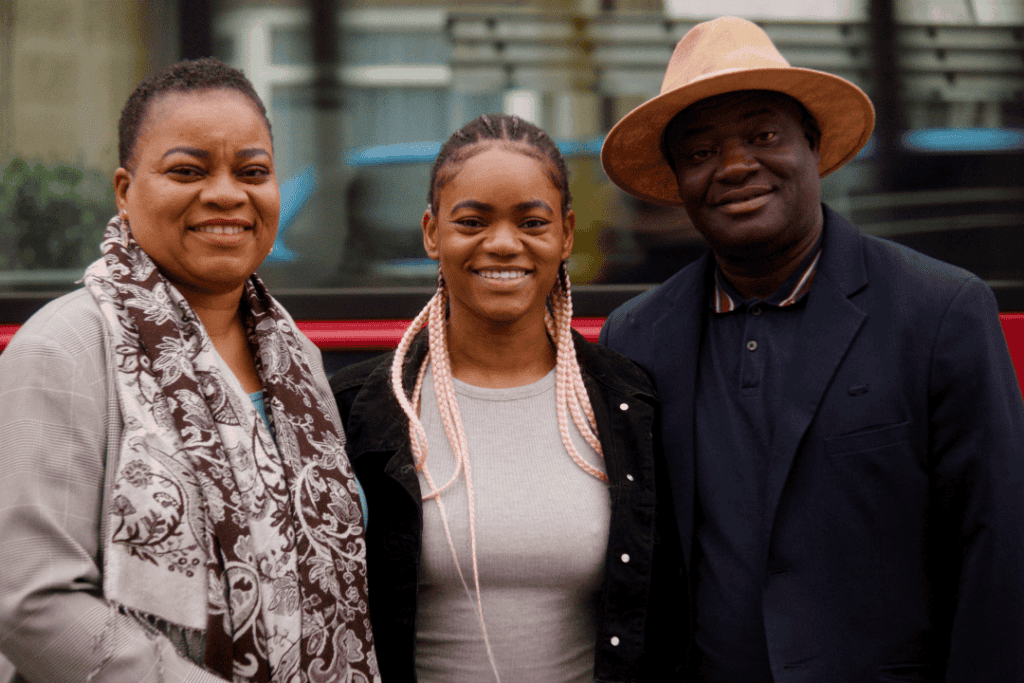 Sarah stands outside with her mother on the right and her father on the left. They're all smiling at the camera. Sarah wears a hearing aid. Her father wears a hat.