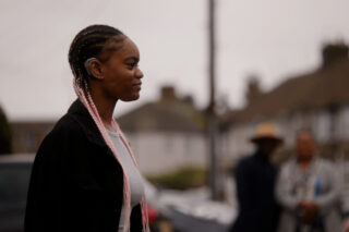 A young woman stands outside, facing sideways to the camera. Her assistive hearing technology is visible at her ear. Her pink braids are over her shoulders. There are people in the background, blurred.