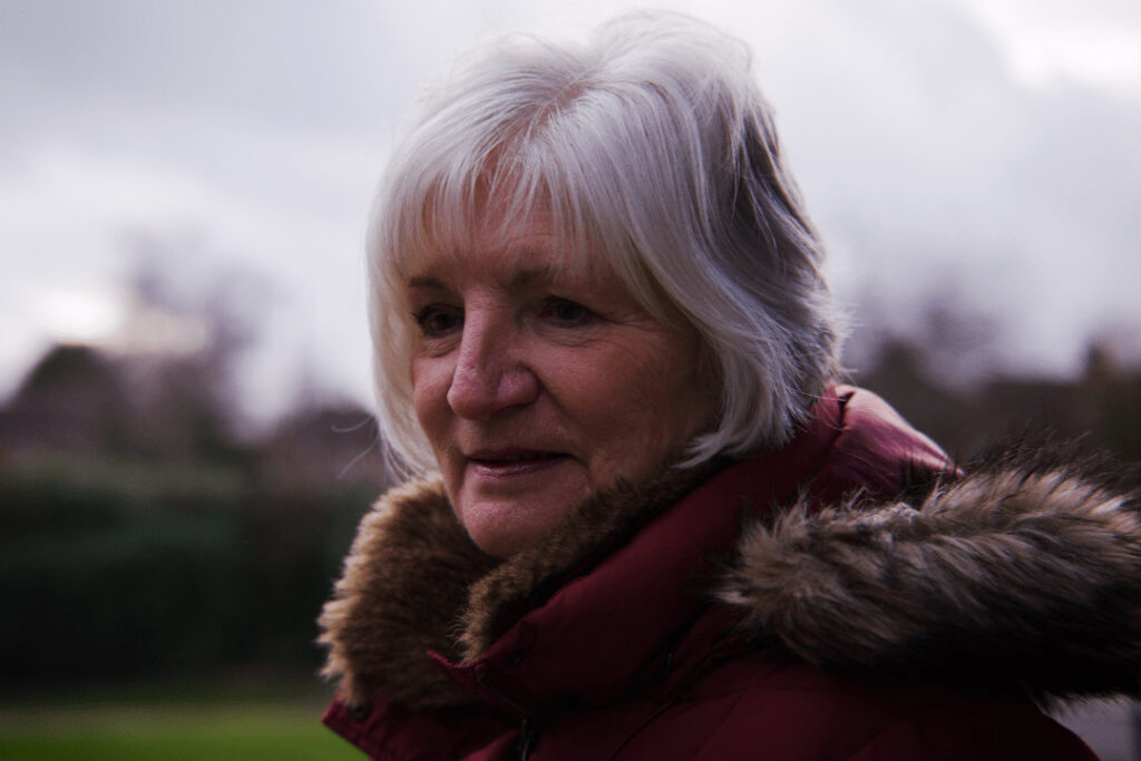 Sheila stands outside in her coat, looking aside from the camera.