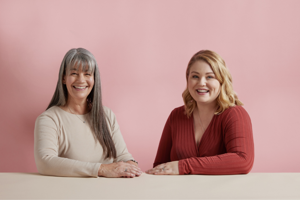 Two women, one older and one younger, sit at a desk in front of a pink background, smiling at the camera.