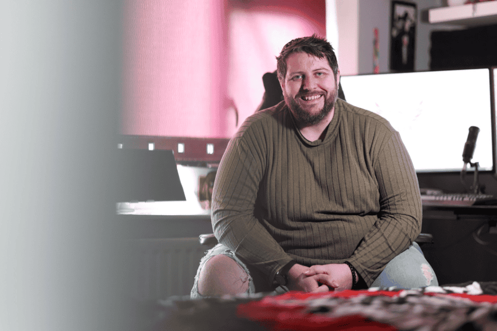 David sits in his computer chair, smiling at the camera with his hands interlaced in his lap. In the background is his computer.