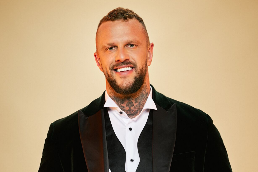 Deaf contributor to Married at First Sight UK 2024, Ross McCarthy. He stands in a tux with his shirt collar open at the neck. He smiles at the camera.