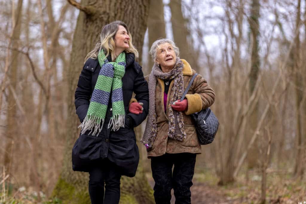 Audrey and Jodie wear winter coats and scarves, walking through a forest and linked arm in arm. They look up into the trees.