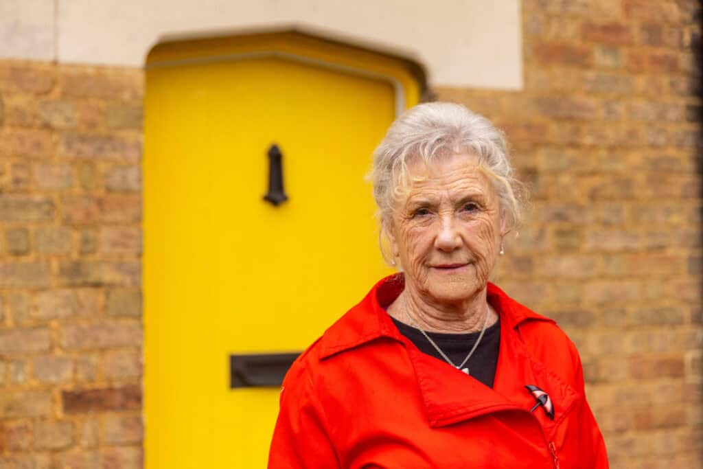 Audrey stands outside of her front door, wearing a red coat and looking seriously into the camera.