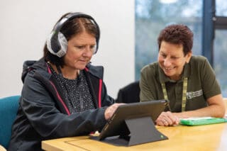 An RNID Near You volunteer helps a member of the public take a hearing test. They sit at a table and use a laptop.