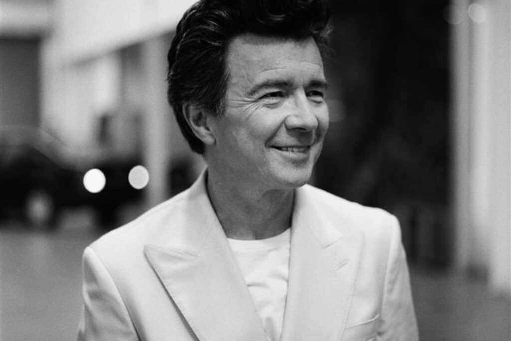 Black and white photo of Rick Astley. He wears a t-shirt under a blazer and looks to the side, smiling.