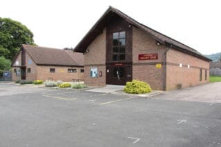 A photo taken outside of Shurdington Community Hall. It's a small building with a carpark.