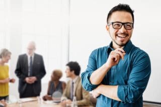 A smiling business man standing in front of a group of people sat around a meeting desk.