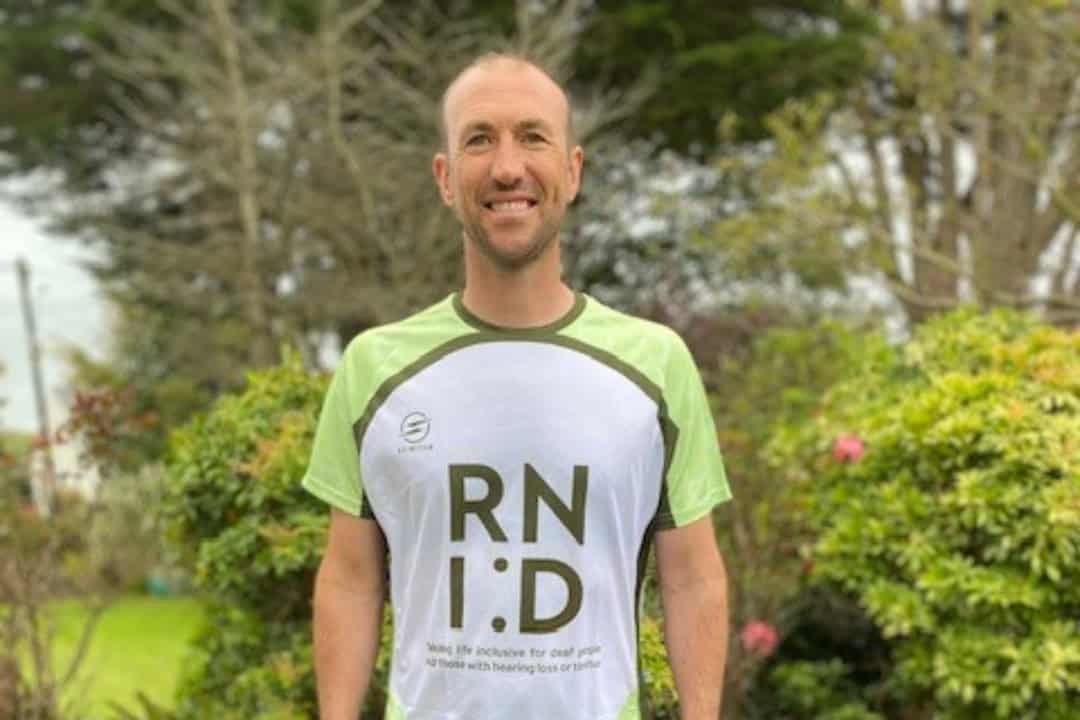 Simon, a tall slim man, is standing outside and wearing an RNID fundraising t-shirt. He smiles for the camera.