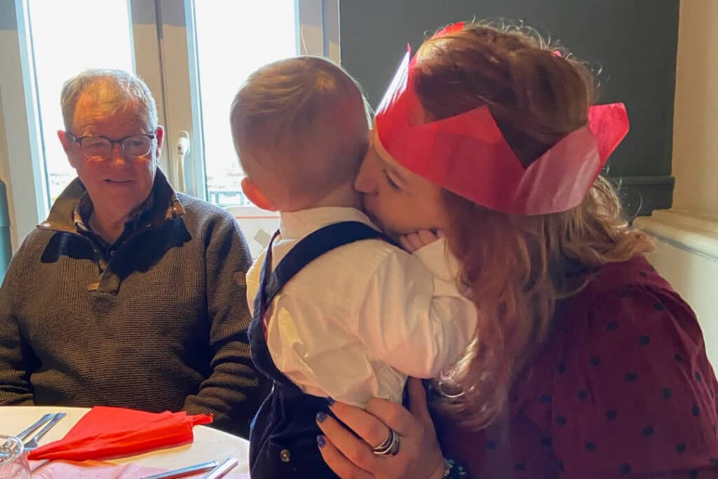 A woman wearing a paper Christmas hat cuddles a child who is sitting on the table. An older man looks at them affectionately. 