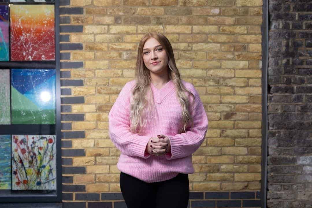 Caris L wears a pink jumper and stands in front of a brick wall, meeting the eye of the camera. 