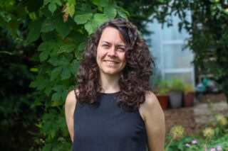 Charlotte, a woman with curly brown hair, stands beside a tree.
