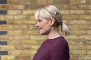 A woman with short blonde hair stands in front of a brick wall. She has a cochlear implant.