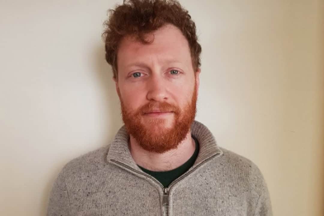A headshot of a man with a beard wearing a beige jumper.