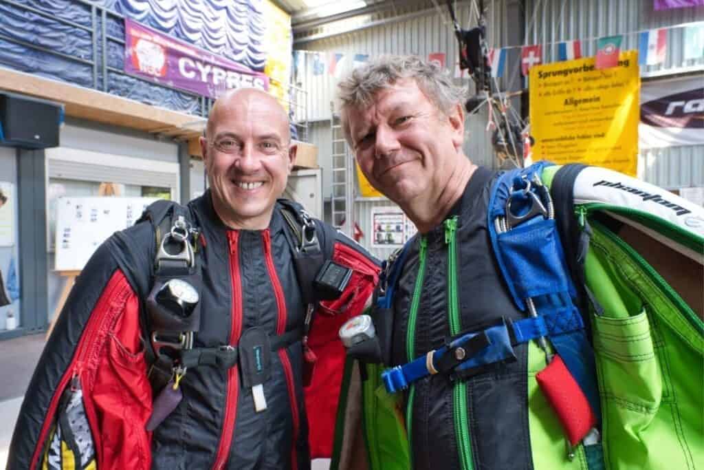 Two smiling men wearing jumpsuits and harnesses ready to skydive.