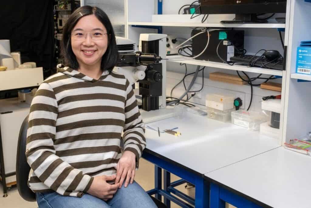 A smiling woman in a striped top and jeans sit at a desk in a laboratory.
