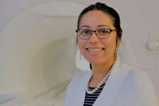 Professor Fatima Hussain wears glasses and a white lab coat. She smiles at the camera.