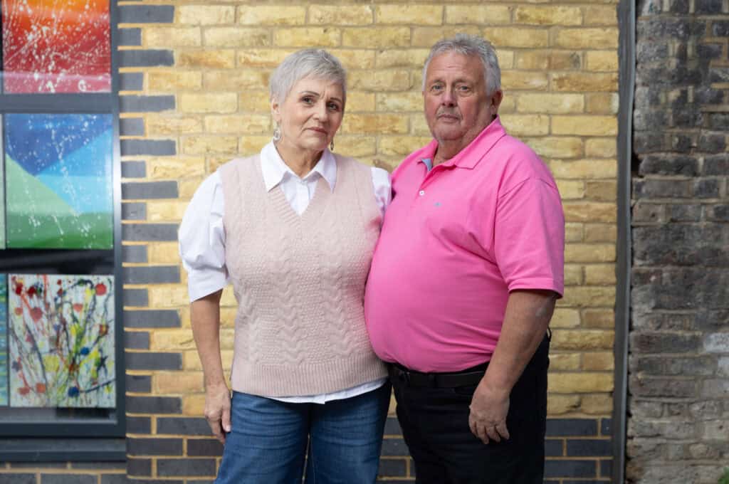 RNID storyteller, Dougie, and his wife, Sue, stand in front of a brick wall. Sue wears a blouse and jeans, and Dougie wear a pink t-shirt and black trousers. 