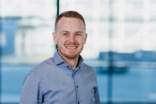 A headshot of RNID-funded scientist, Wouter. He wears a blue shirt and stands in front of a large glass window, smiling.