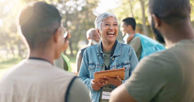 A woman laughing holding a clipboard and engaging with a group of people outdoors