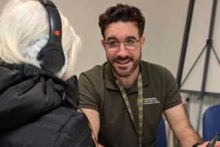 A man with brown hair and beard wears a green RNID Near You t-shirt and helps someone take a hearing check.