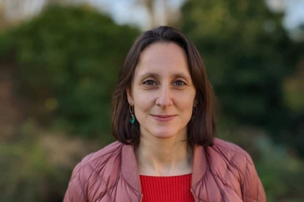 A woman stands outside. She has shoulder length brown hair wearing a pink jacket smiles.
