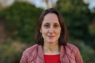 A woman stands outside. She has shoulder length brown hair wearing a pink jacket smiles.
