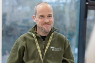 RNID Near You, Wayne, wears a green fleece and lanyard and smiles.
